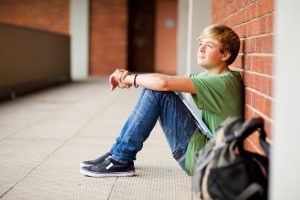 Young male student with backpack sits against a brick wall and looks into the distance