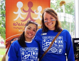 Two smiling women wearing blue Ride for Refuge shirts stand in front of a Welcome Home Refugee House banner