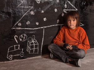 boy in red shirt sits next to picture of Christmas tree drawn on a chalk board