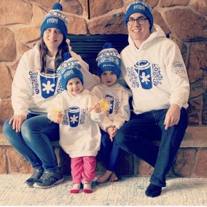 Brian Ellis, his wife and two small daughters sit in front of a fireplace while wearing CNOY toques.