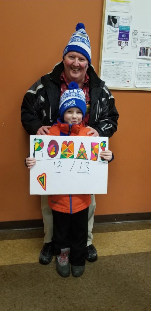 A young boy and an older man wearing winter coats and hats hold a sign that reads 12 or 13 Romans