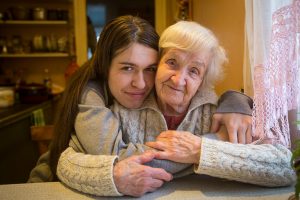 A young woman cuddles an elderly woman as they sit at a kitchen table