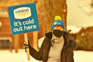 Woman wearing a mask and toque holds a Coldest Night sign