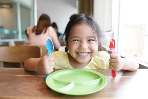 Little girl smiles as she holds she holds a colourful spoon and fork