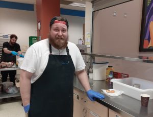 A burly young man with a beard and wearing an apron stands next to an counter in an industrial kitchen