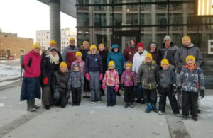 A group of smiling propel wearing yellow toques stand outside a building