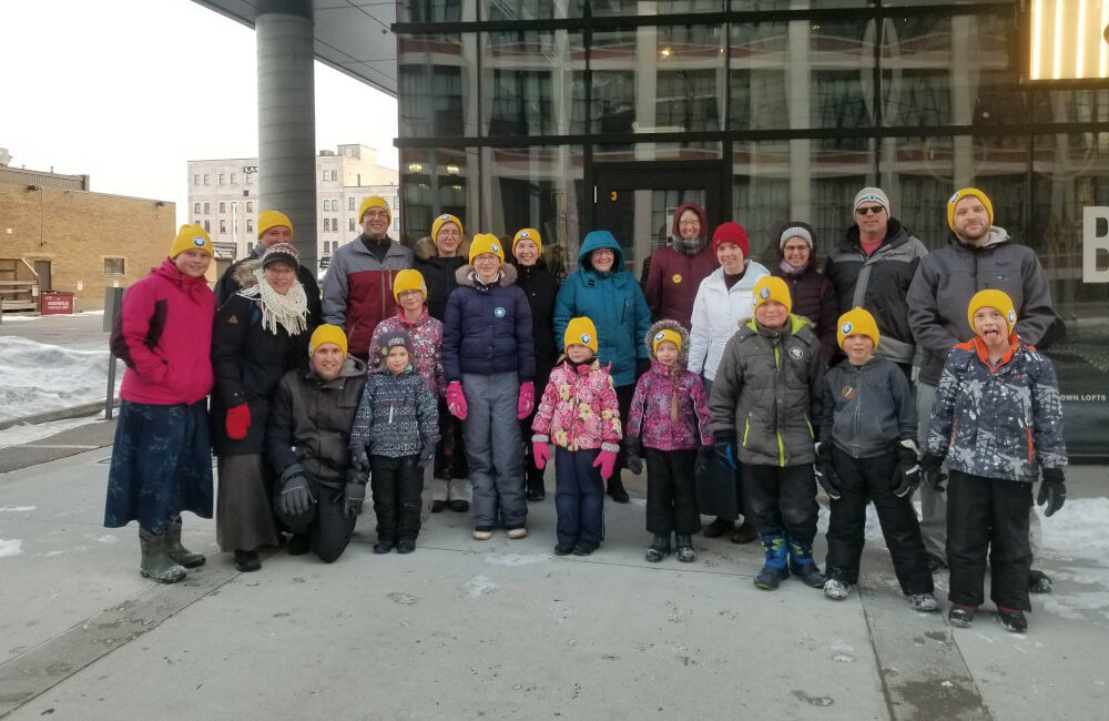 A group of smiling people wearing winter coats and yellow toques stand outside a building