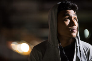 Young Asian man wearing a hoodie looks up toward a light on a dark street