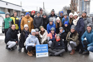 A group of people wreaking winter coats and toques pose with a Coldest Night of the Year sign