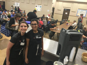 Two female volunteers wearing aprons smile as they stand in Ray of Hope's dining room