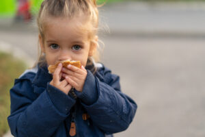 A small blonde girl looks at the camera with sad eyes as she eats a piece of bread