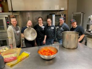 Six smiling women stand behind a steel kitchen counter