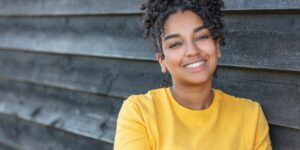 A young Black woman in a yellow Tshirt smiles as she leans against a dark wooden wall